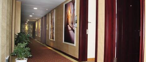 A hotel corridor with framed pictures on the walls, a potted plant, and a red carpet.