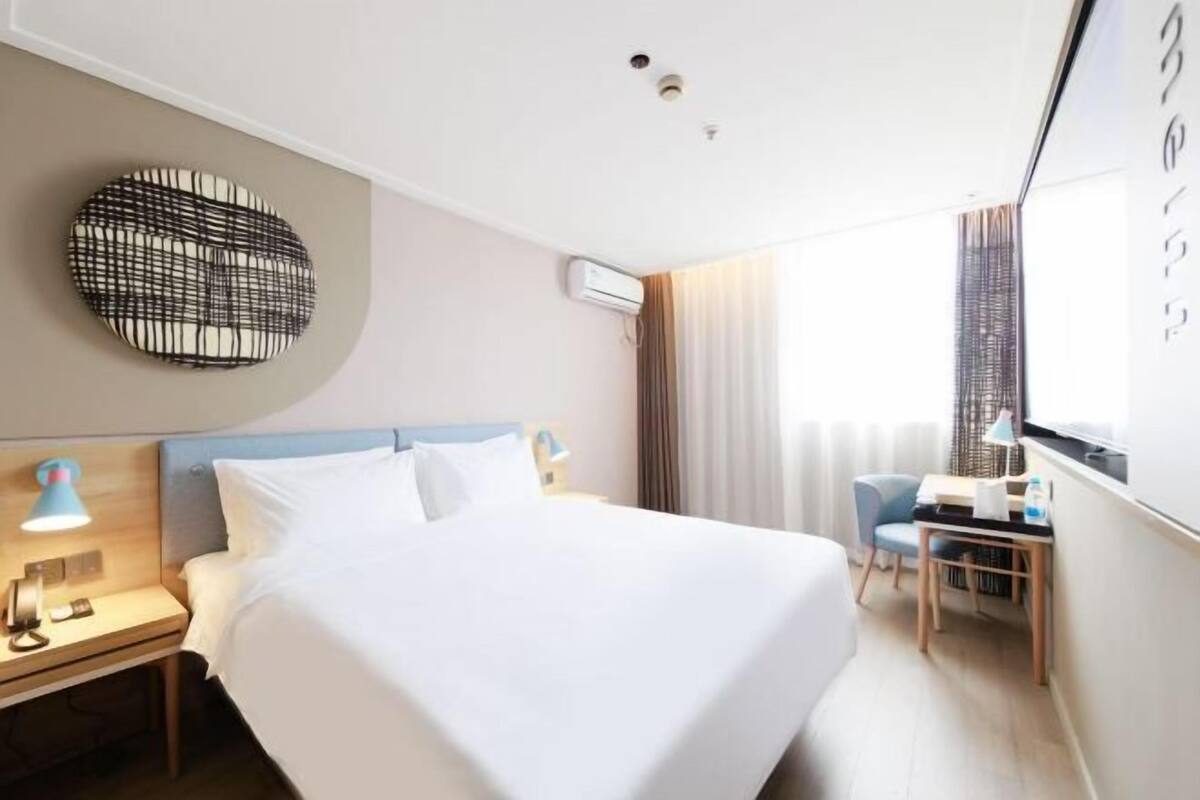 A hotel room with a bed, bedside table, lamp, desk, chair, and a window with curtains.