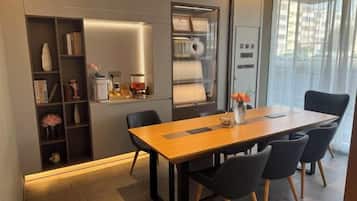 A modern dining area with a wooden table, black chairs, a built-in bookshelf, and a kitchen area with a refrigerator and oven.