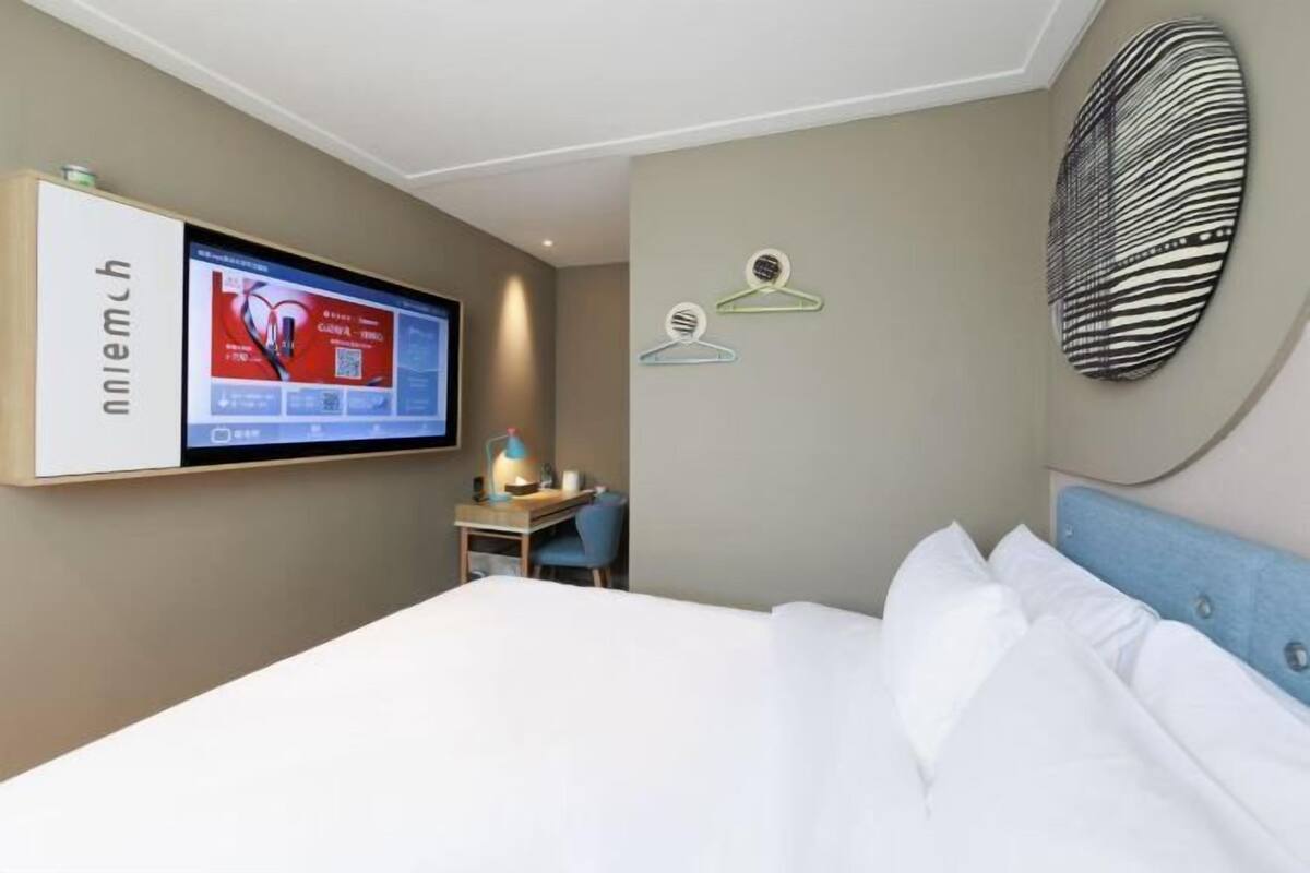 A hotel room with a bed, a desk with a chair, a TV mounted on the wall, and a wall-mounted coat rack.
