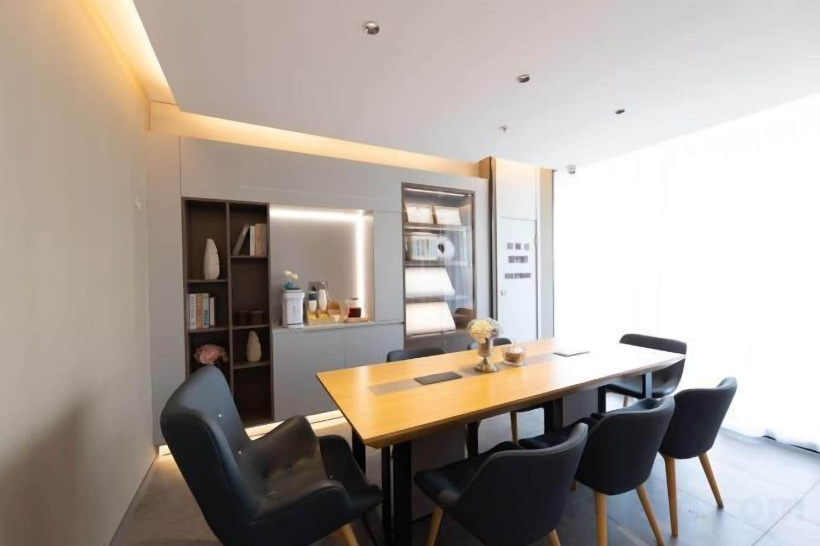 A modern dining area with a wooden table and black chairs, a built-in bookshelf, and a kitchen area with a refrigerator and cabinets.