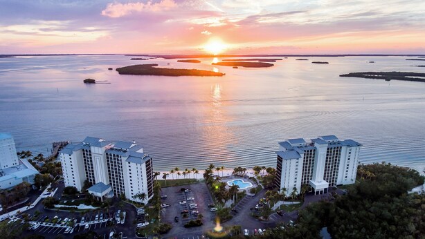 A high-rise hotel with multiple floors, a parking lot, and a view of the sunset over a body of water with several islands.