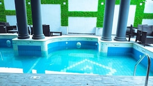 Ein Schwimmbecken mit gefliestem Sonnendeck, Liegestühlen und einer grünen Wand mit geometrischem Muster.