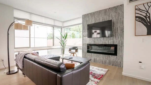 Un salon moderne avec un canapé en cuir, un téléviseur à écran plat fixé sur un mur à texture, et une grande fenêtre laissant entrer la lumière naturelle.