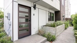 Un bâtiment moderne doté d’une grande porte vitrée, d’un bac en béton planté de végétaux et d’un chemin menant à l’entrée.