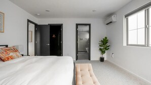 Une chambre à coucher moderne avec un grand lit, un banc, une plante en pot et une salle de bain visible à travers une porte ouverte.