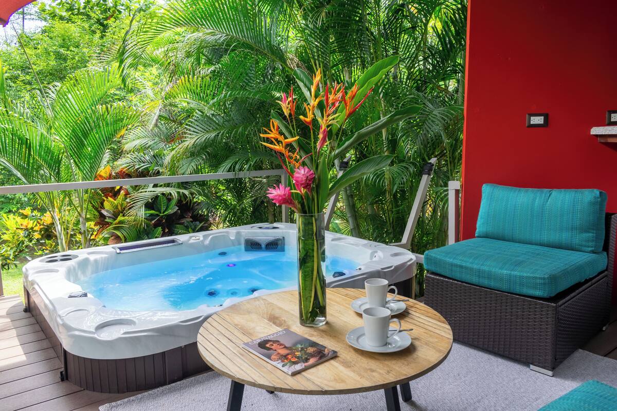 A hot tub on a deck with a wicker chair and a table with a magazine.