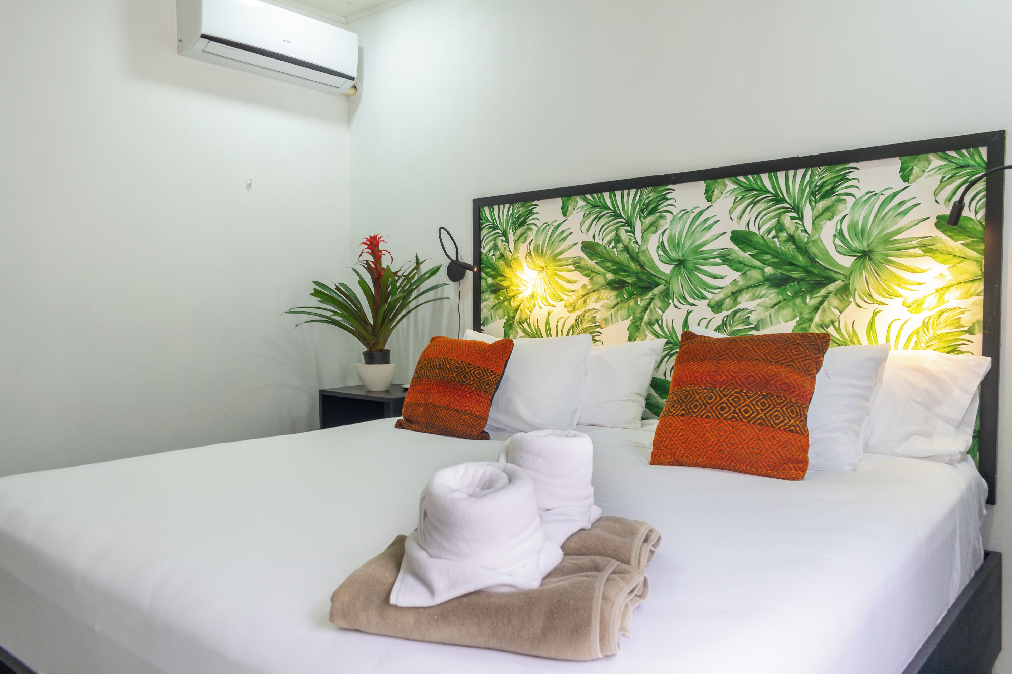 A hotel room with a bed, white linens, two orange patterned pillows, a headboard with a tropical leaf design, and a potted plant on the nightstand.