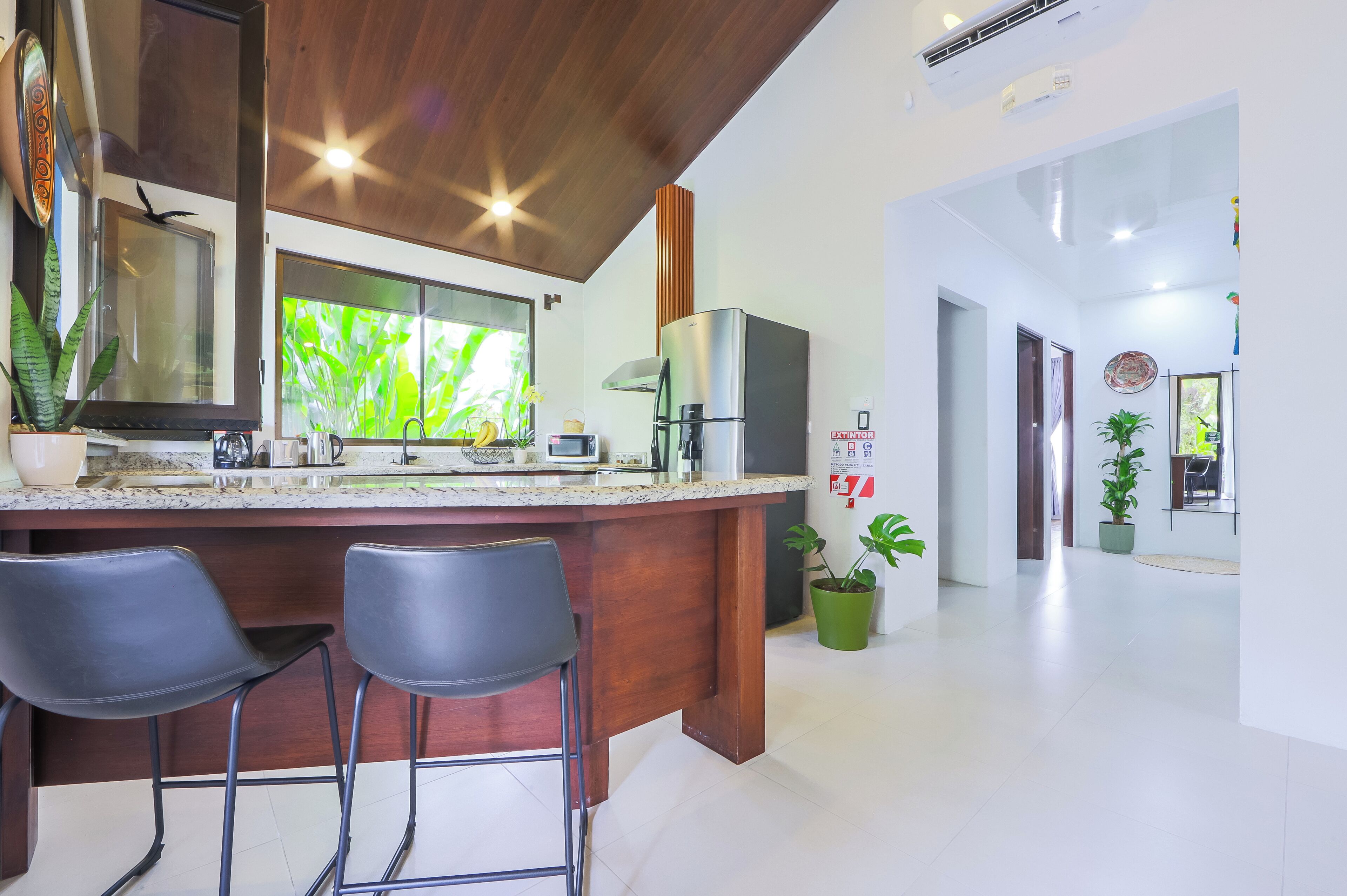 A modern kitchen with a wooden island, two bar stools, a large window with a view, and a stainless steel refrigerator.