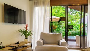 A room with a TV mounted on the wall, a sofa, a small table with a plant, and a sliding glass door leading to an outdoor area with greenery. - Bukala Village Lodge (Peñas Blancas)