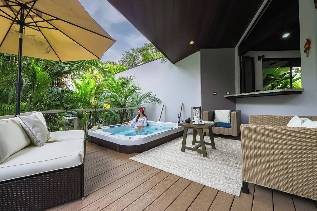 A modern outdoor living area with a hot tub, wicker furniture, and a view of lush greenery.