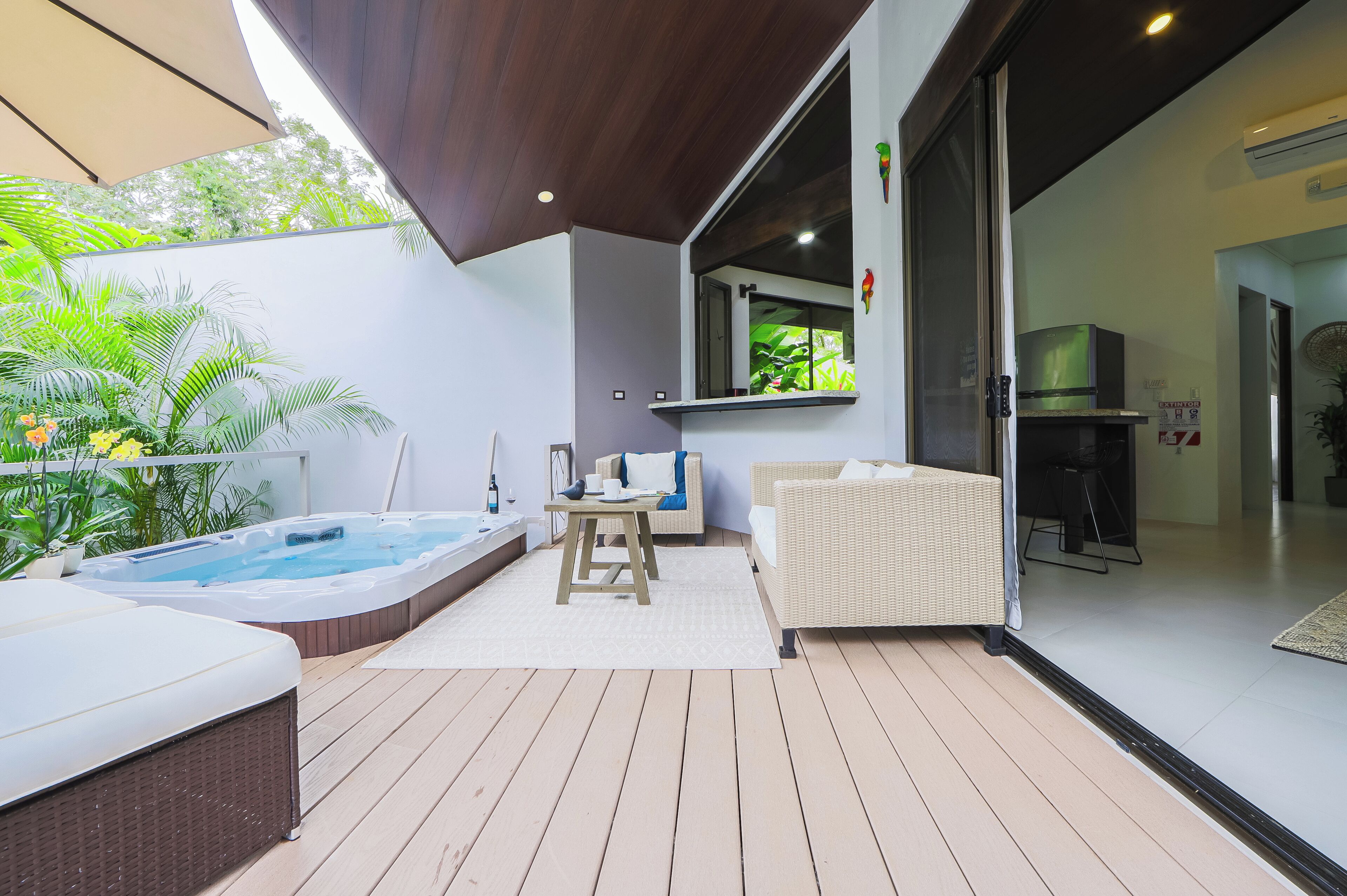 A modern outdoor patio with a hot tub, wicker furniture, and a view of the greenery.