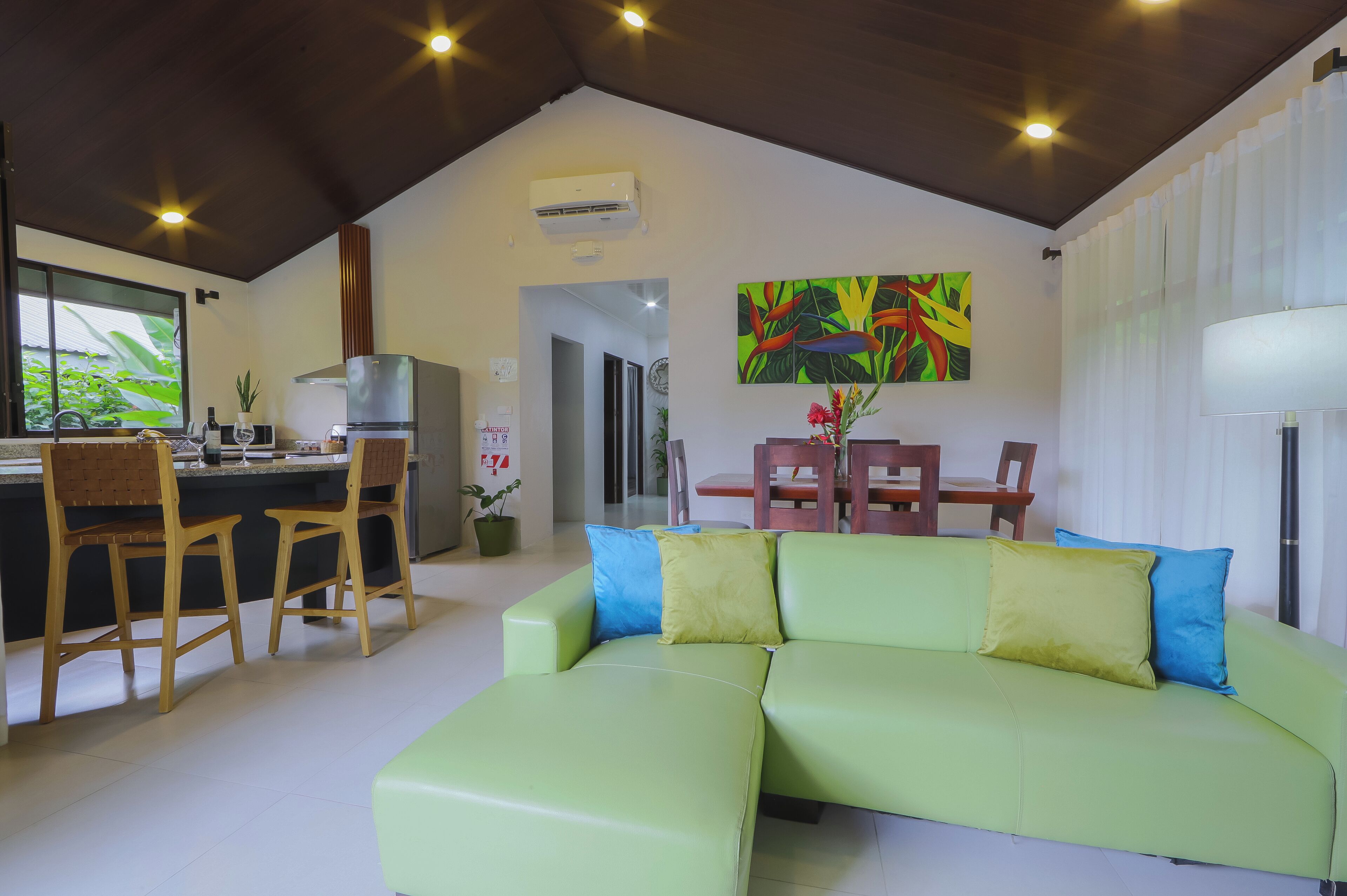 Una cocina moderna con zona de comedor, un sofá verde con cojines azules y amarillos, y un cuadro abstracto y colorido en la pared.