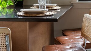 Un coin repas avec une table dressée pour deux personnes, une plante en pot et une fenêtre avec des stores.