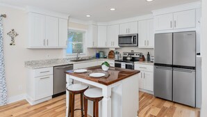 A modern kitchen with white cabinets, a stainless steel refrigerator, a microwave, and a wooden island with stools. - Luxurious & Comfy Haven: Prime Location ~ Pool ~ Hot Tub ~ Fire Pit ~ Gym ~ Pkg! (Wilmington)