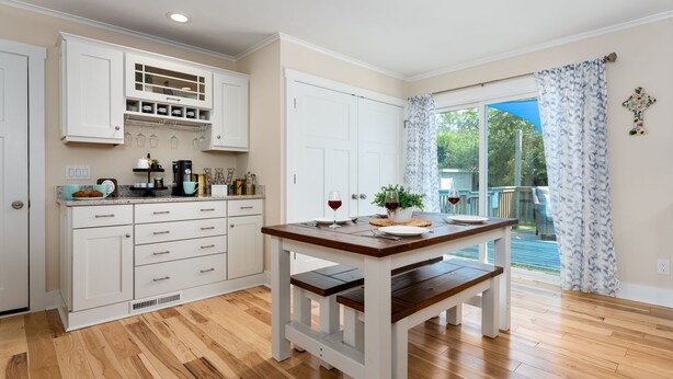 A kitchen with wooden flooring, white cabinets, a dining table with benches, and a view of the outdoors through a glass door. - Luxurious & Comfy Haven: Prime Location ~ Pool ~ Hot Tub ~ Fire Pit ~ Gym ~ Pkg! (Wilmington)