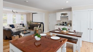 A modern living room with a wooden dining table set for two, a sofa, and a kitchen area with white cabinets and a dining table. - Luxurious & Comfy Haven: Prime Location ~ Pool ~ Hot Tub ~ Fire Pit ~ Gym ~ Pkg! (Wilmington)