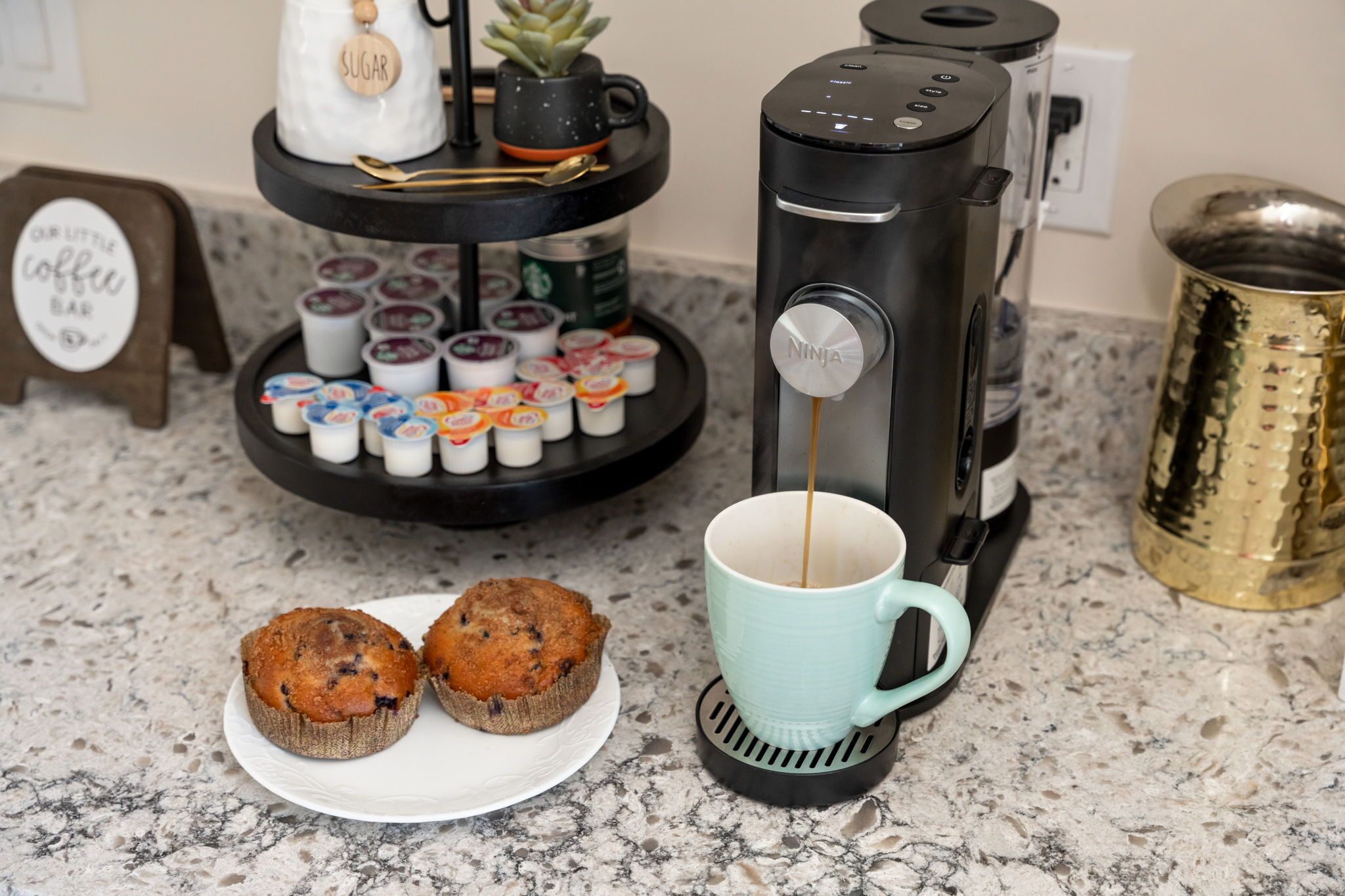 A coffee station with a coffee maker, muffin display, and a sign that reads "OR LITTLE COFFEE BAR".