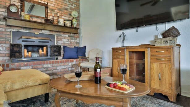 A cozy living room with a brick fireplace, a flat-screen TV, a wooden table with a bottle of wine and glasses, and a fruit platter. - Maxine's Place- A Wonderful Family Legacy (Abilene)