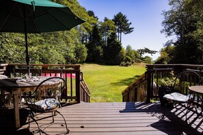 A wooden deck with a table and chairs, a green umbrella, and a view of a grassy area with trees.