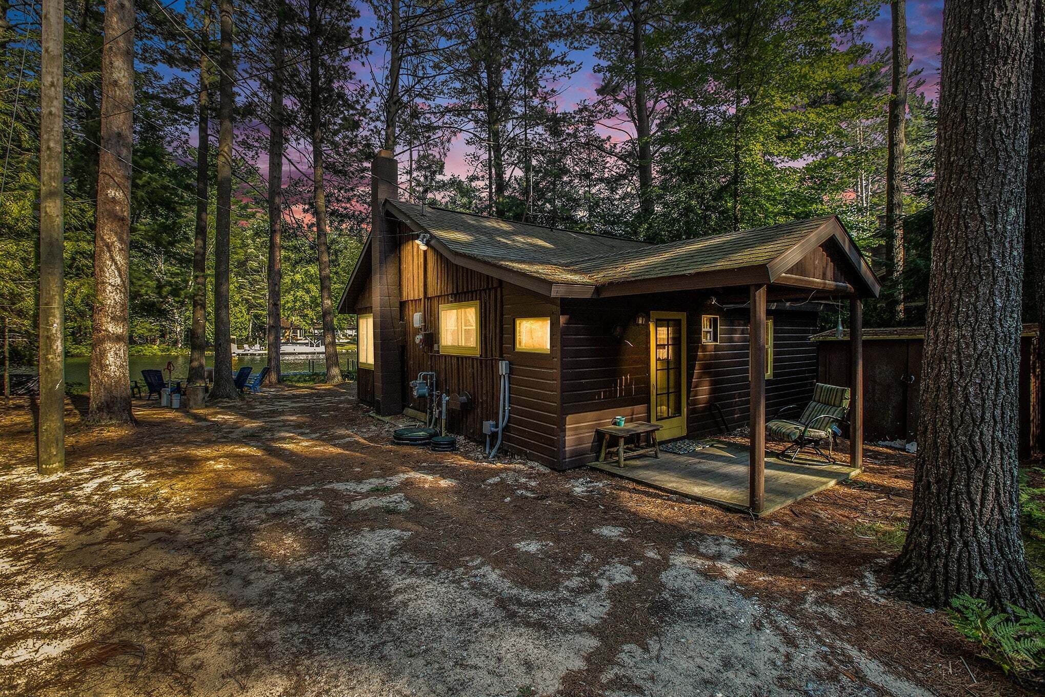 Un chalet rustique doté d’une véranda, entouré de grands arbres et bordé d’un sol en terre battue.