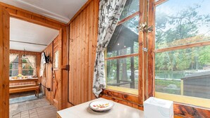 Interior de una cabaña de madera con comedor, un ventanal con vista a un lago y una mesa con un plato y una caja.