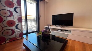 Ett modernt vardagsrum med en stor platt-TV placerad på en vit TV-bänk, ett glasbord och utsikt över en balkong med mönstrade gardiner.