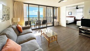 A spacious living room with a large sofa, a glass coffee table, a TV, and a balcony with a view of the city. - Ocean-view lanai, 1 block to Waikiki Beach (Honolulu)