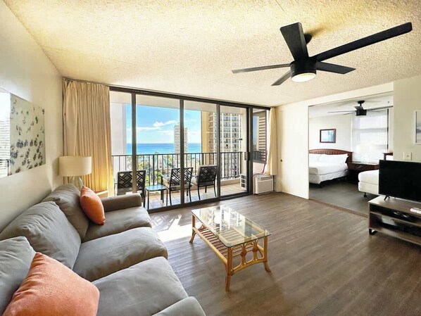 A spacious living room with a large sofa, a glass coffee table, a TV, and a balcony with a view of the city.