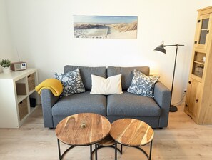 A grey sofa with floral cushions, two wooden side tables, a radio, and a bookshelf.