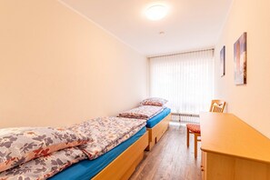 Ein kleiner, gut beleuchteter Raum mit zwei Einzelbetten, einem Holztisch und einem Stuhl.