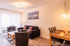 A modern living room with a sofa, chairs, a dining table, and a cityscape painting on the wall. - Komfortable, Ruhige und Zentral Gelegene Ferienwohnung Seeschwalbe auf Borkum - Strandstraße 5-7 (Borkum)