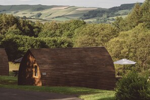 A row of wooden cabins with sloped roofs, surrounded by lush greenery and hills in the background. - Hot Tub Cabin With Views Over The Brecon Beacons (Brecon)