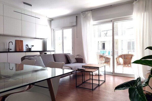 A modern living space with a glass dining table, a grey sofa, wicker chairs, and a large window with curtains.