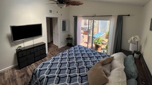 Un dormitorio con cama, televisor, ventilador de techo, puerta corrediza de vidrio a un patio y una pequeña mesa con una planta.