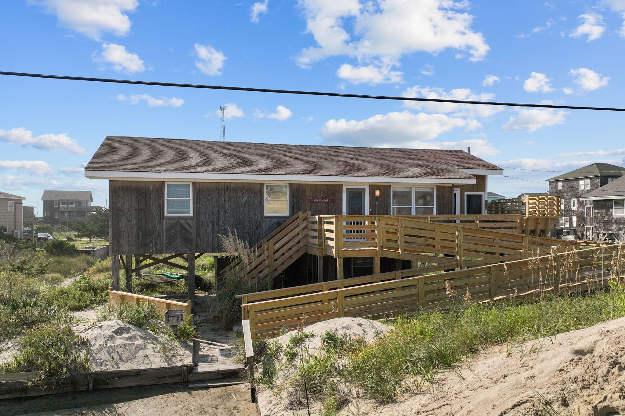 A wooden beach house with a staircase leading to a deck, surrounded by sand and vegetation.