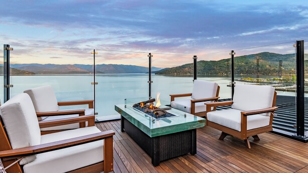 A deck with wooden furniture and a glass-top table with a fire feature, overlooking a body of water and distant mountains. - Waterfront Luxe Seasons ~8~ Boat Slip (Sandpoint)