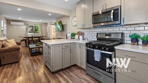 A modern kitchen with grey cabinets, a stainless steel microwave, and a gas stove. There is a dining area with a table and chairs, and a living room with a sofa and a TV. - Two-Story Midtown Home—Newly Renovated, Fire Pit, and Peaceful Outdoor Space for Relaxation! (Coeur D Alene)