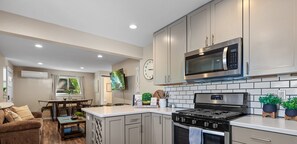A modern kitchen with grey cabinets, a stainless steel microwave, and a gas stove. There is a dining area with a table and chairs, and a living room with a sofa and a TV.