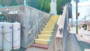 A staircase with yellow and green tiles leading up to a concrete wall with a chain-link fence and gas cylinders. - Hotel Frontier (Isinomaki)