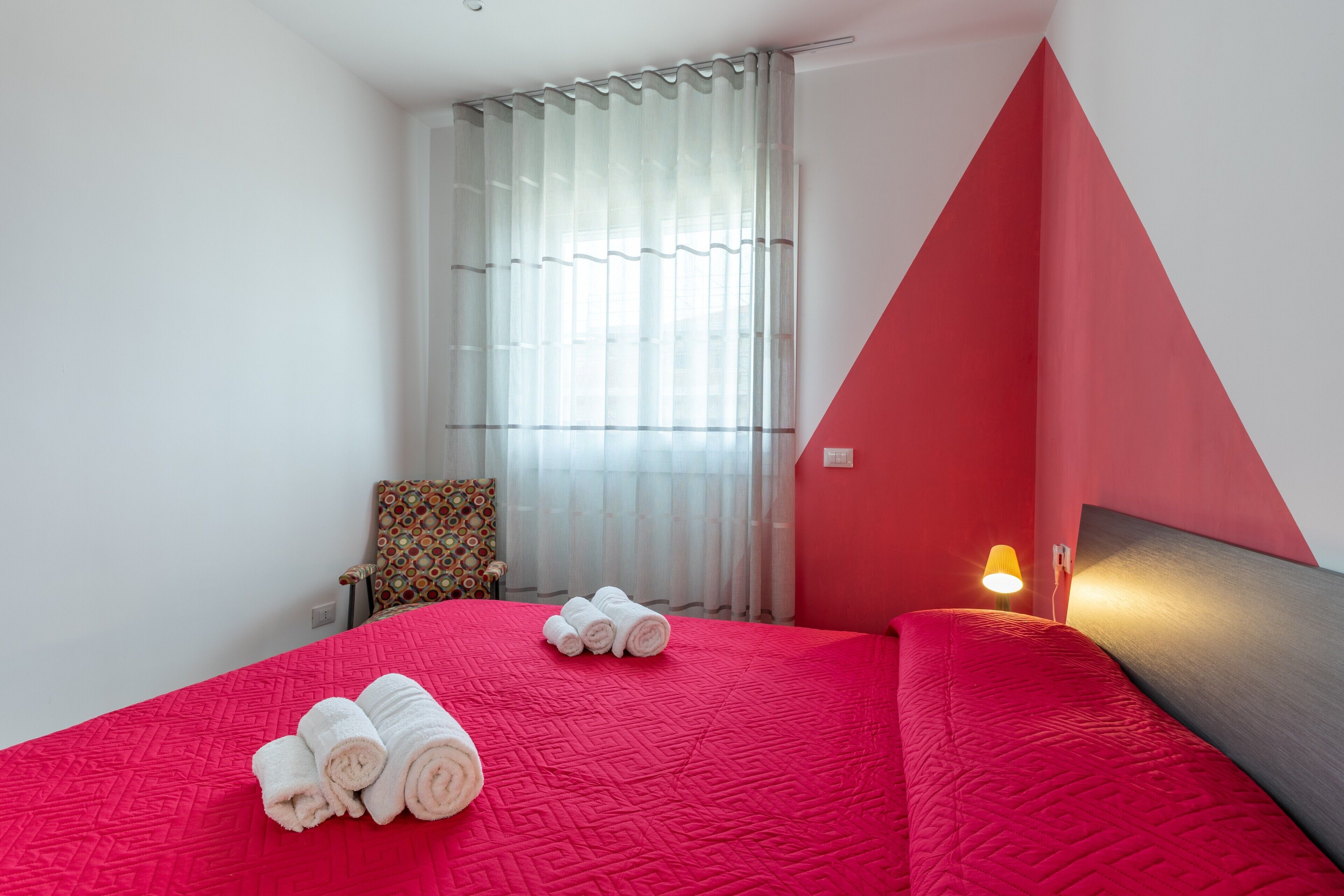 A hotel room with a red bedspread, a chair with a patterned cushion, a window with white curtains, and a bedside lamp.