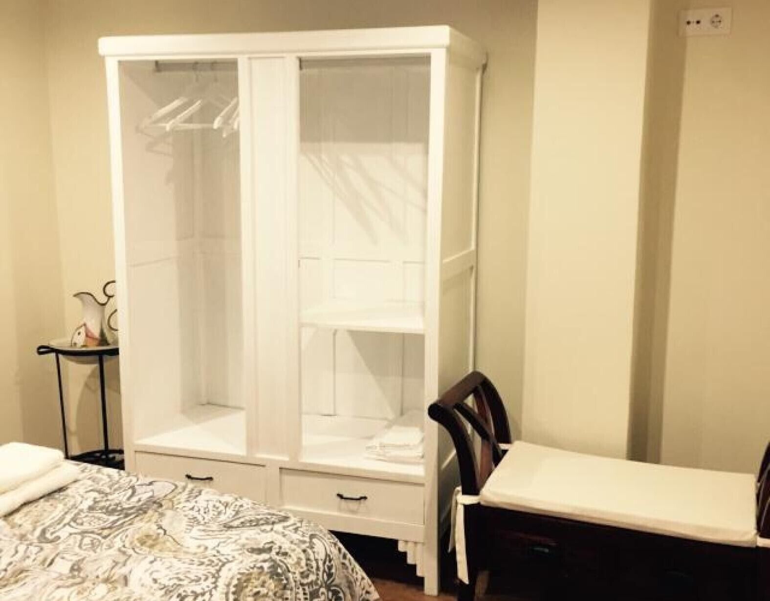 A bedroom with a large white wardrobe, a bed with patterned bedding, and a small bedside table.