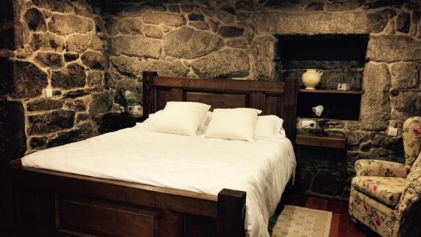Un dormitorio con pared de piedra, cama de madera, mesita de noche, una silla y una estantería con objetos.