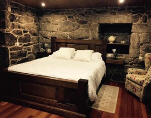 Un dormitorio con pared de piedra, cama de madera, mesita de noche, una silla y una estantería con objetos.