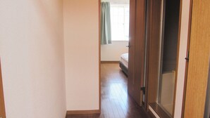 Ένας στενός διάδρομος με ξύλινο δάπεδο οδηγεί σε ένα υπνοδωμάτιο με ένα κρεβάτι και ένα παράθυρο με κουρτίνες.
