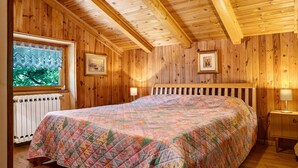 Un dormitorio con paneles de madera, una cama, un ventanal con cortinas de encaje, un radiador y cuadros enmarcados en las paredes.