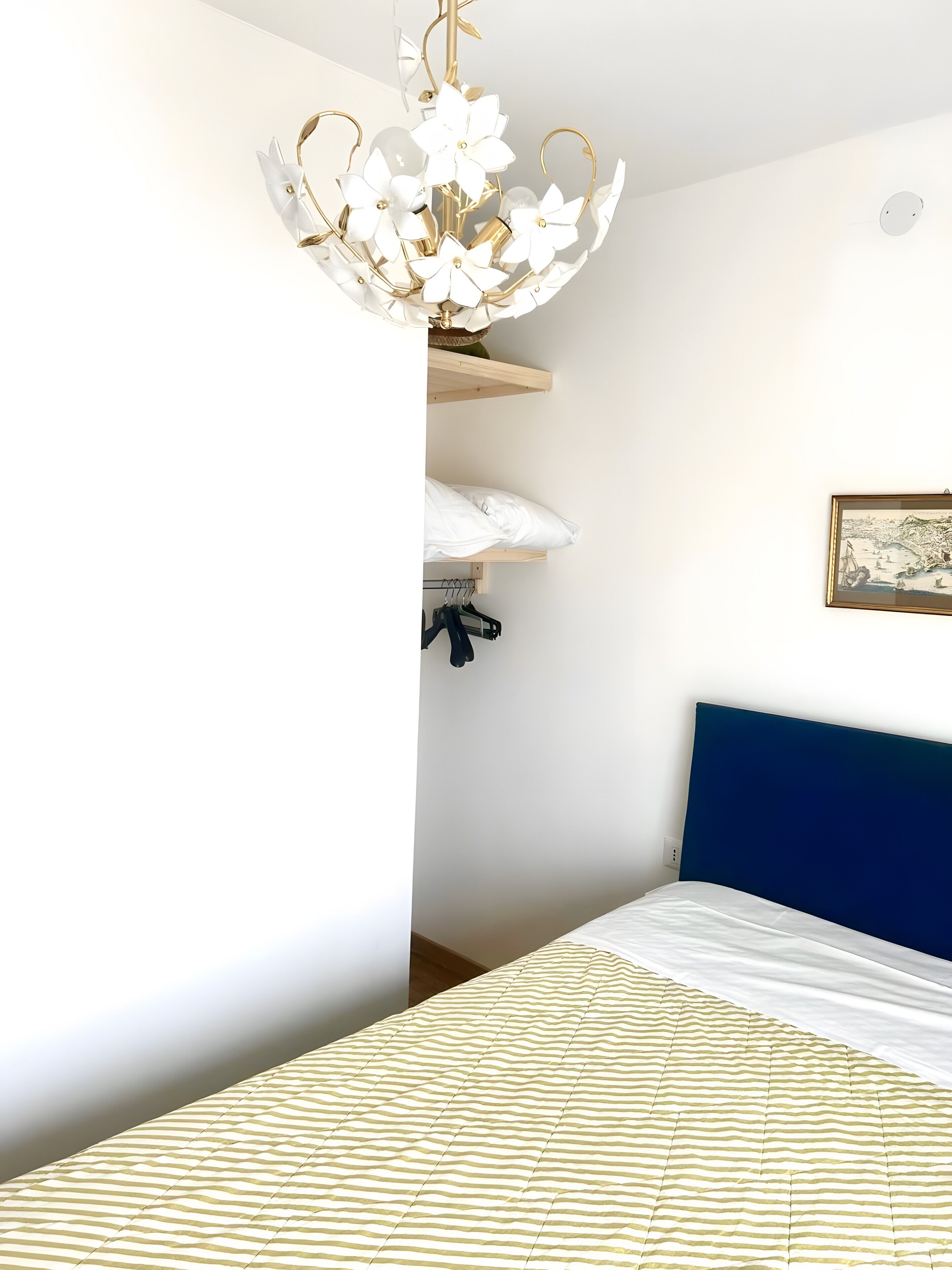 A bedroom with a bed, a blue headboard, a chandelier, and a wall-mounted shelf with clothes.