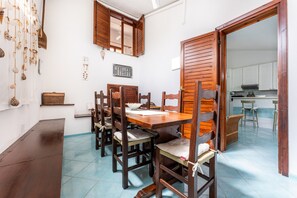 A dining area with wooden furniture, a kitchen visible through an open door, and a tiled floor.