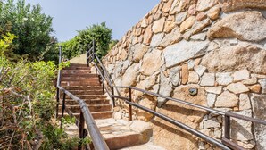 Un escalier en pierre, agrémenté de rampes métalliques, mène au sommet d’une colline, le tout étant entouré de verdure.