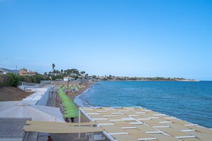 Un hôtel situé en bord de mer, avec de nombreuses chaises longues et des parasols alignés, une vue dégagée sur la mer et, au loin, une côte parsemée de bâtiments.